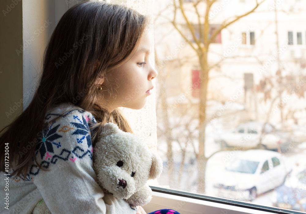 Sad little girl holds a teddy bear looks out the window. Stock