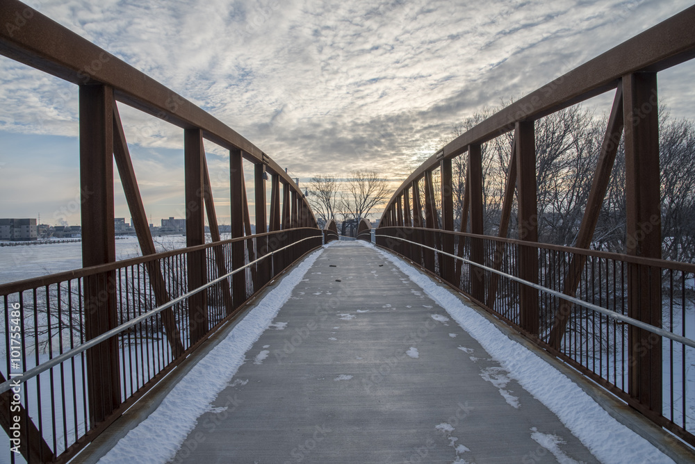 A walking bridge spans a river and leads to a snowy winter path.