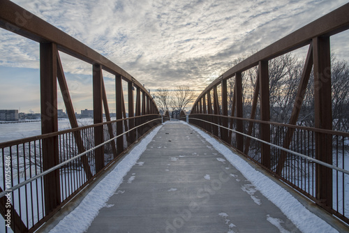 A walking bridge spans a river and leads to a snowy winter path.