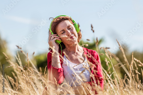 outdoors relaxation - gorgeous middle aged woman enjoying listening to music in headphones in dry high grass, summer daylight