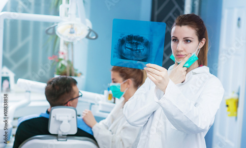 Dentist looking at roentgen of human jaw. Patient with another doctor are blurred on chair in background.