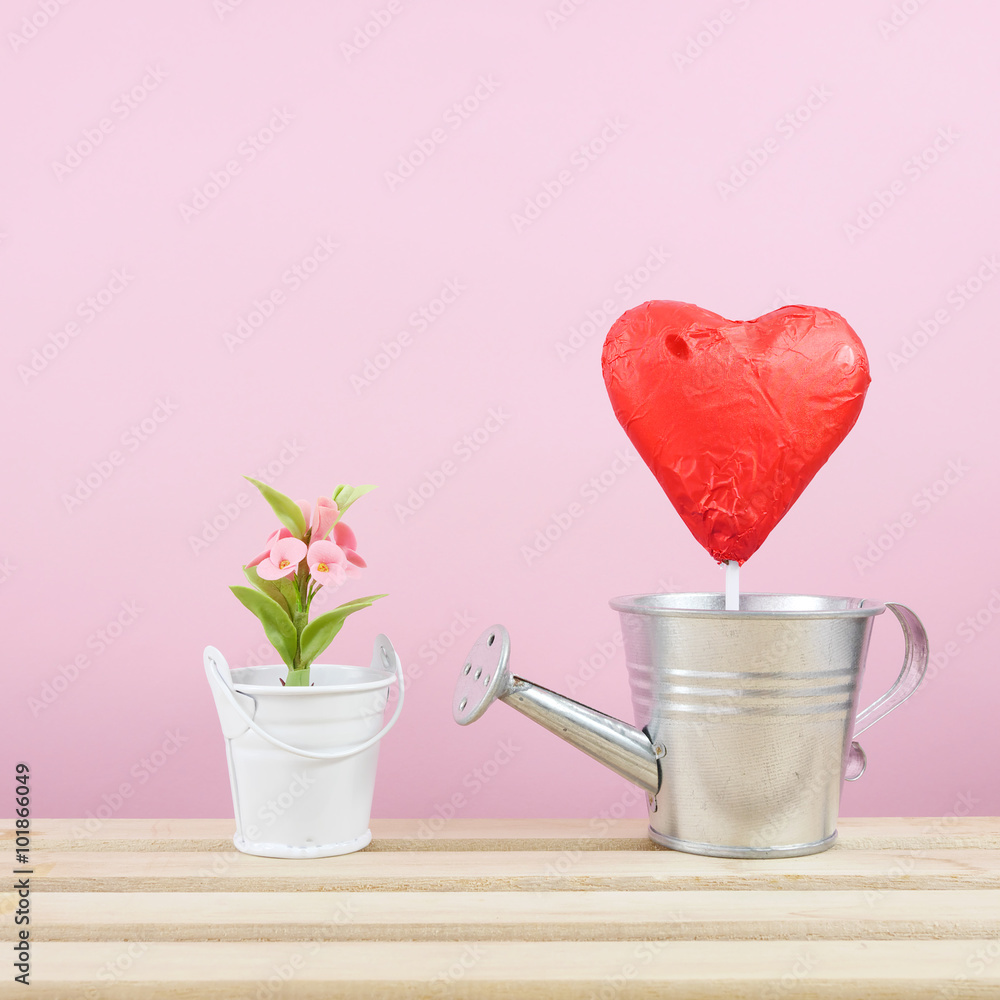 The red foiled chocolate heart stick with small silver watering can and mini fake flower in small bucket on wooden tray for Valentine's day. The red foiled chocolate heart stick with small silver watering can and mini fake flower in small bucket on wooden tray for Valentine's day.