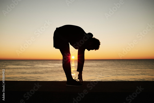 Young muscular build man doing stretching exercise on the beautiful sea background during sunset, attractive athlete doing workout outdoors at sunrise, fitness and healthy lifestyle concept.