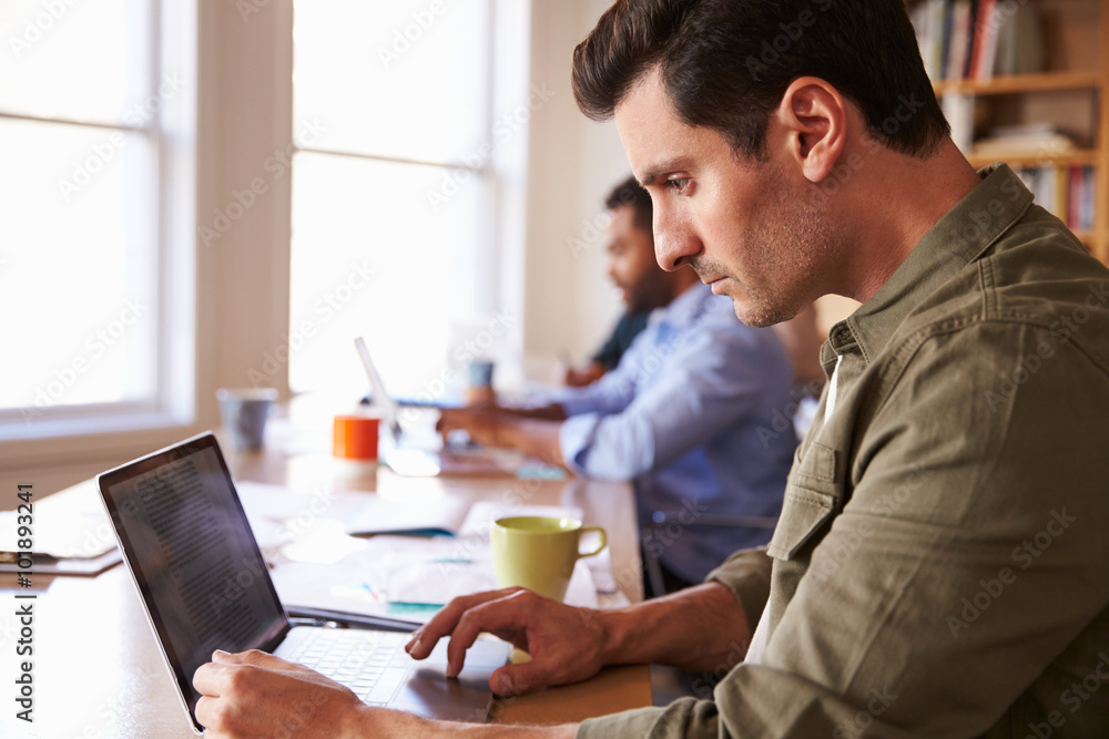 Businessman Using Laptop At Desk In Busy Office Businessman Using Laptop At Desk In Busy Office