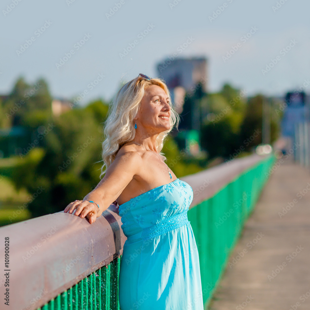 Portrait of positive elderly blonde woman on the bridge. Mature beautiful blonde in blue dress walks on the bridge. the concept of transition to old age