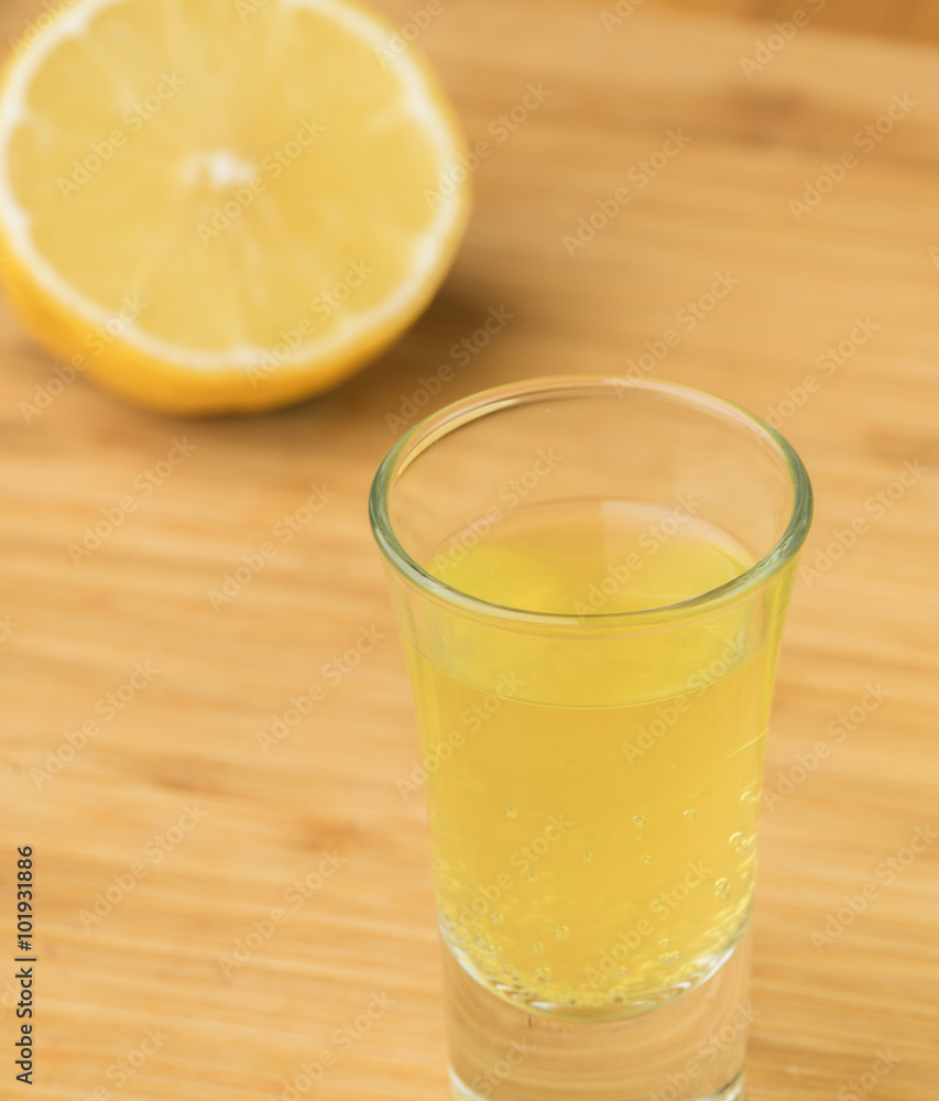 Limoncello/Lemon-flavored liqueur from Southern Italy made with zest of Femminello St. Teresa lemons shown on light wood surface with shot glass and cut fresh lemon Limoncello/Lemon-flavored liqueur from Southern Italy made with zest of Femminello St. Teresa lemons shown on light wood surface with shot glass and cut fresh lemon