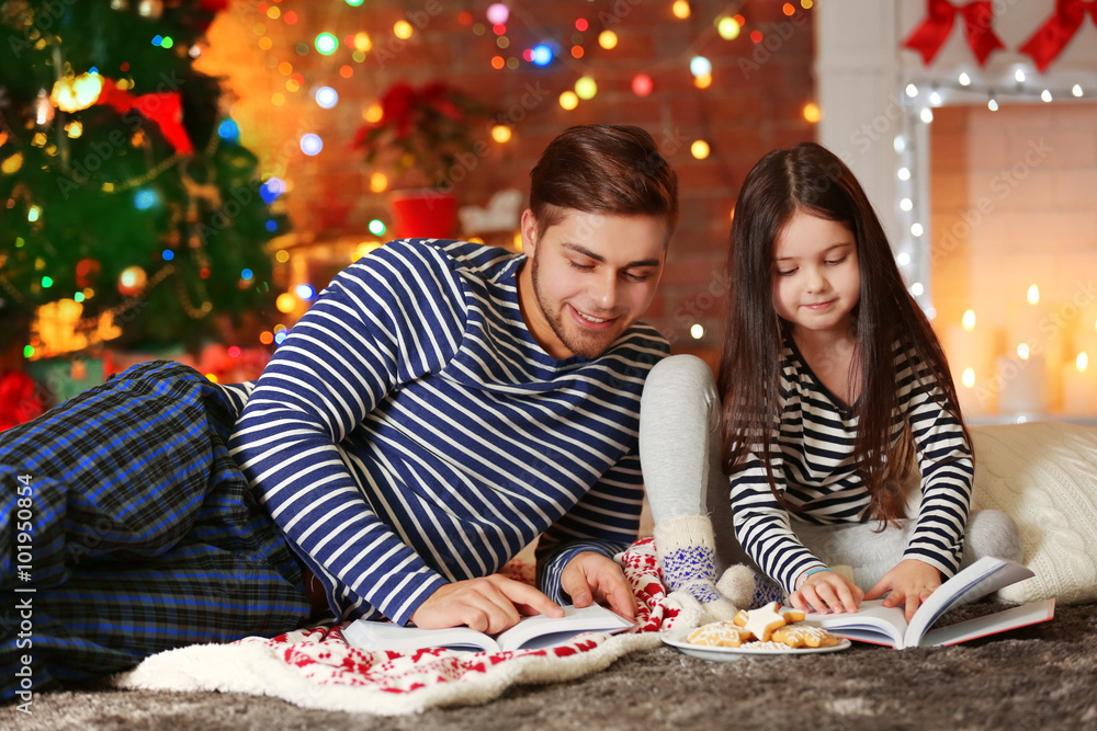 Fototapeta premium Older brother with little sister reading books and eating cookies in Christmas living room