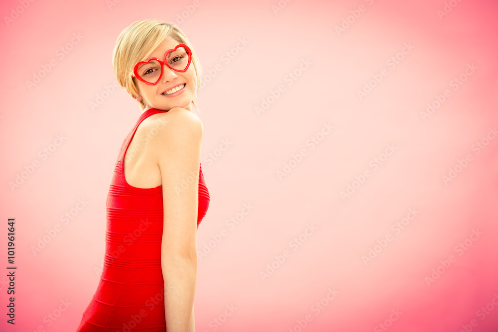 Young beautiful woman with hearth shaped glasses in red dress shy smile on pink for valentine day