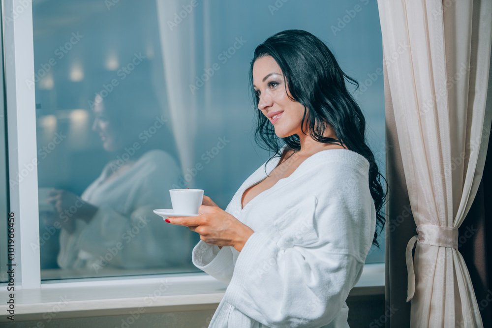 Fototapeta premium Getting warm with fresh coffee. Beautiful young woman in white bathrobe drinking coffee and looking through a window