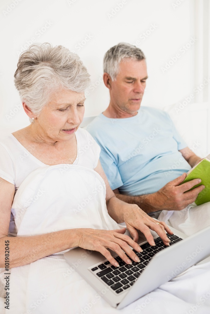 Relaxed senior couple at bed using laptop and reading book