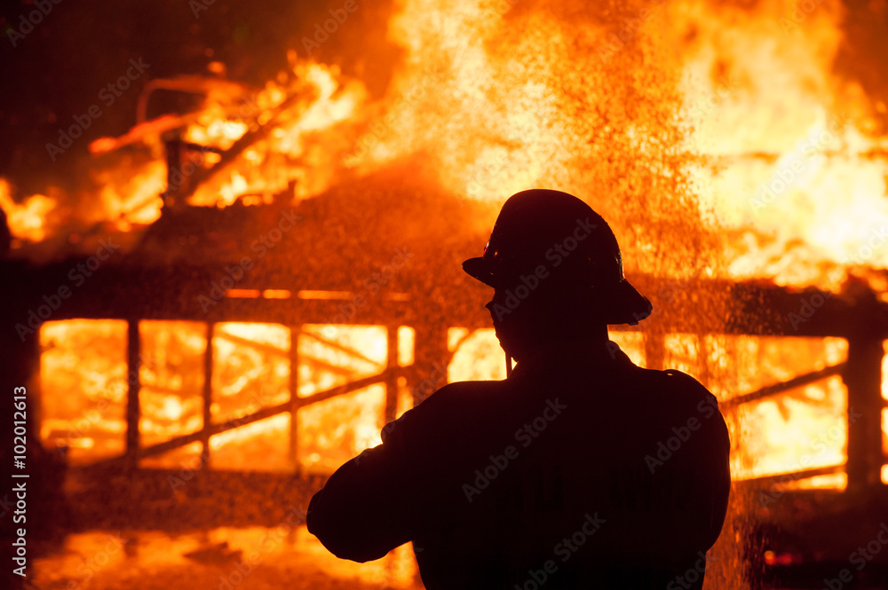 Fototapeta premium Silhouette of Firemen fighting a raging fire with huge flames of burning timber