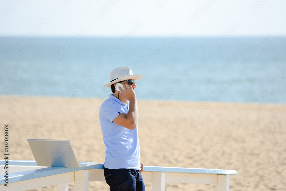 Busy male working on the laptop and talking on mobile. Man in hat speaking on smartphone over blue sky and ocean beach 