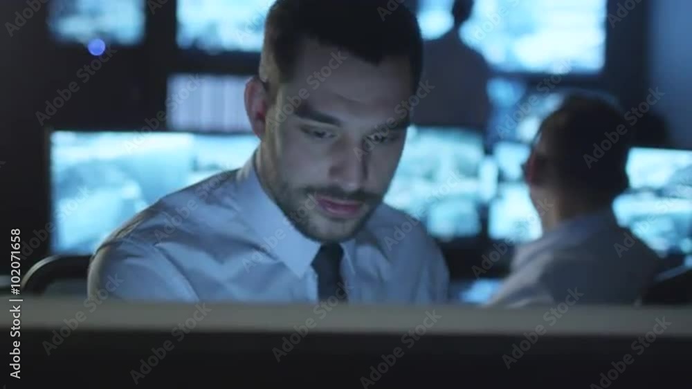 Happy technical support specialist is drinking coffee while working on a computer in a dark monitoring room filled with display screens. Shot on RED Cinema Camera. Happy technical support specialist is drinking coffee while working on a computer in a dark monitoring room filled with display screens. Shot on RED Cinema Camera.