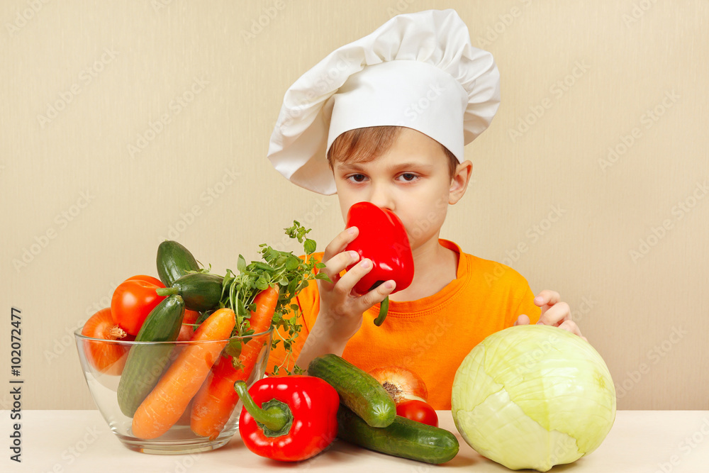Little boy in chefs hat chooses fresh vegetables for salad at the table Little boy in chefs hat chooses fresh vegetables for salad at the table