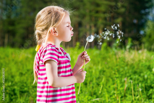 Cute blond little girl blowing a dandelion in summer park on sunny day