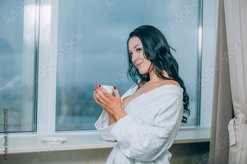 Getting warm with fresh coffee. Beautiful young woman in white bathrobe drinking coffee and looking through a window Free Download Getting warm with fresh coffee. Beautiful young woman in white bathrobe drinking coffee and looking through a window - GFXLoad