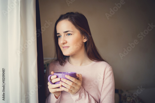 Young woman drinking tea in the morning sitting by the window.Woman drinking coffee in sunshine sitting near window in sun light enjoying her morning coffee.