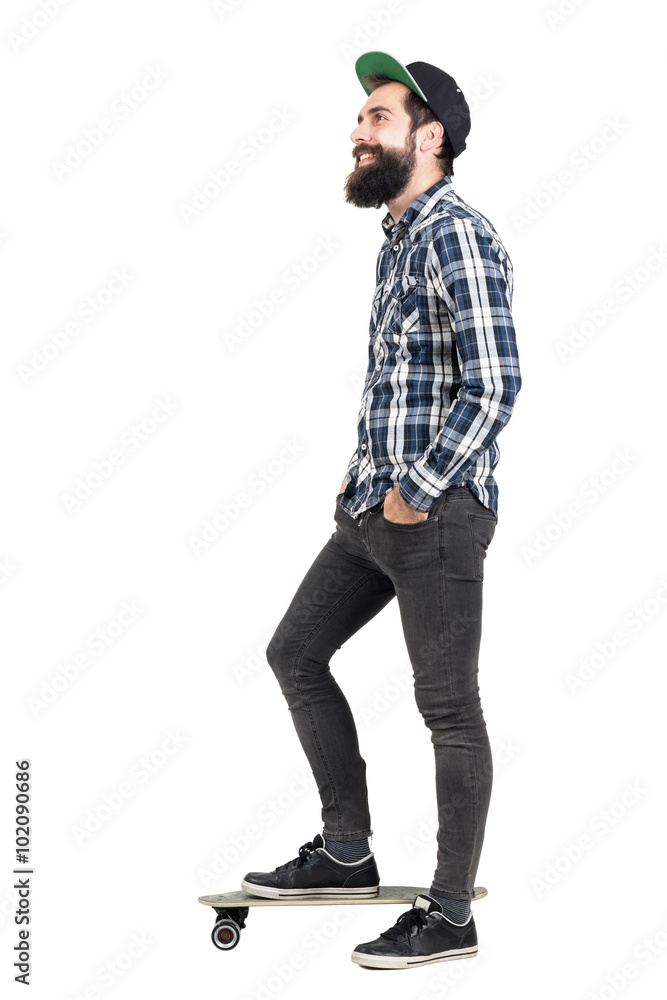 Side view of bearded hipster with cap standing on skateboard looking away smiling. Full body length portrait isolated over white studio background. Side view of bearded hipster with cap standing on skateboard looking away smiling. Full body length portrait isolated over white studio background.