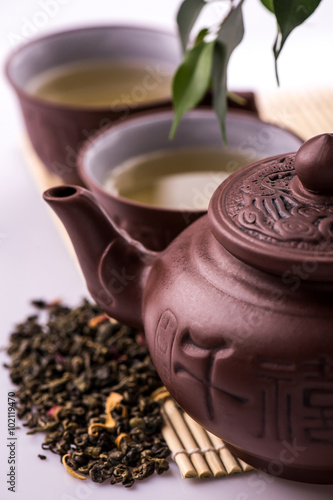 Green tea in a cup and teapot, petals of green tea on a bamboo mat on a white background © nataliiaGL