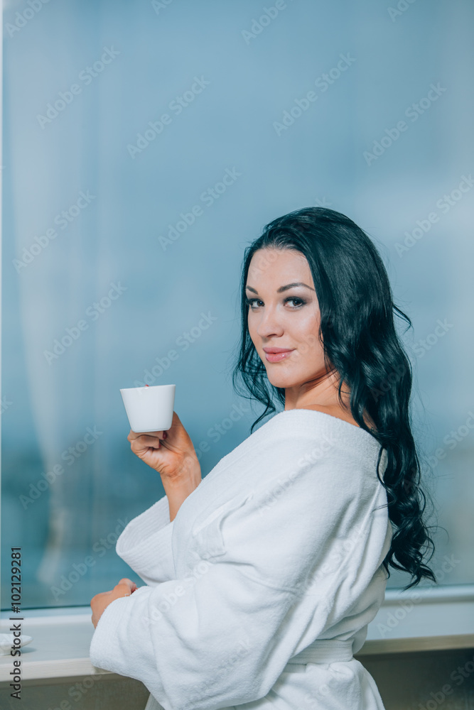 Getting warm with fresh coffee. Beautiful young woman in white bathrobe drinking coffee and looking through a window Getting warm with fresh coffee. Beautiful young woman in white bathrobe drinking coffee and looking through a window