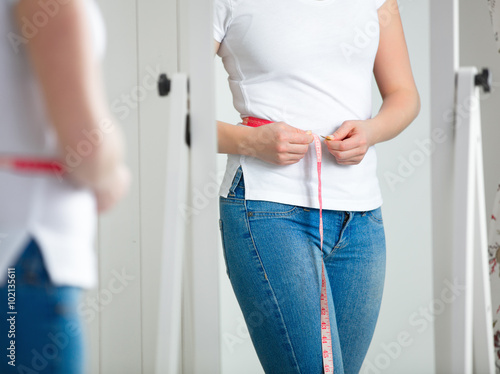 Fotografi young woman measuring waist with measuring tape in front of a mirror