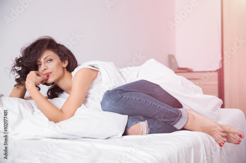 Brunette long hair woman in blue jeans lies on white bed linen