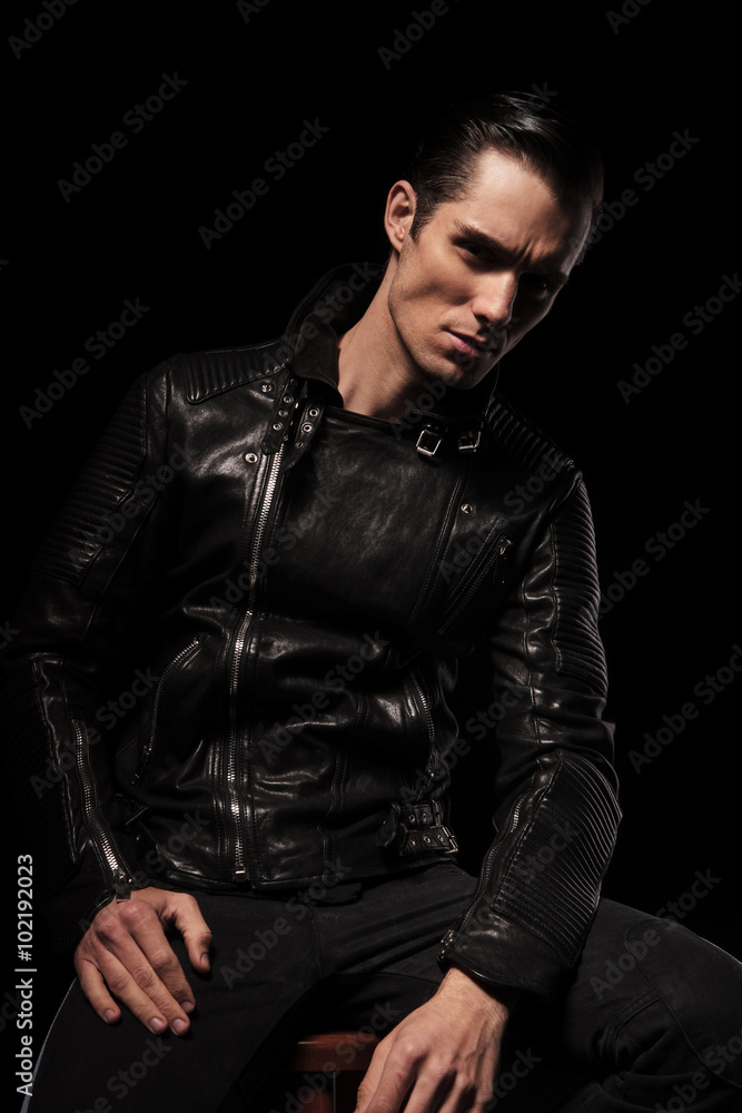 man in black leather jacket posing seated in dark studio backgro