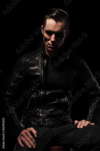 man in black leather jacket posing seated in dark studio