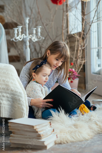 Portrait of a smiling young cute mother and daughter reading a book lying and relax on the floor in a bright big white room next to the window