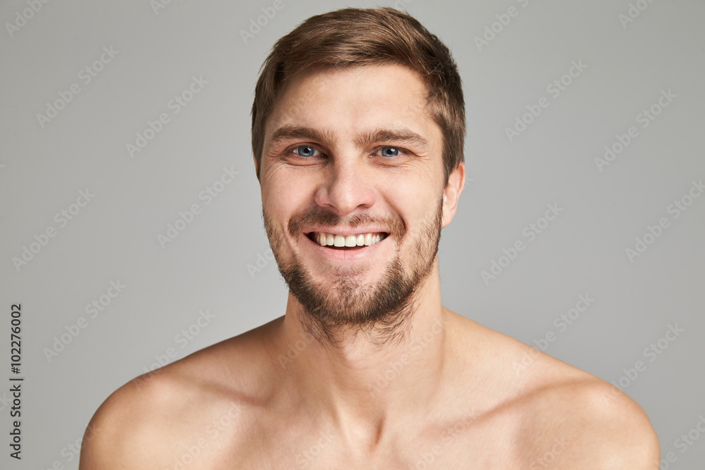 Obraz premium Portrait of a smiling young man with bare swimmers shoulders on a gray background, powerful, beard, charismatic, adult, brutal, athletic, edited photo, bright smile, white teeth smile, look in camera
