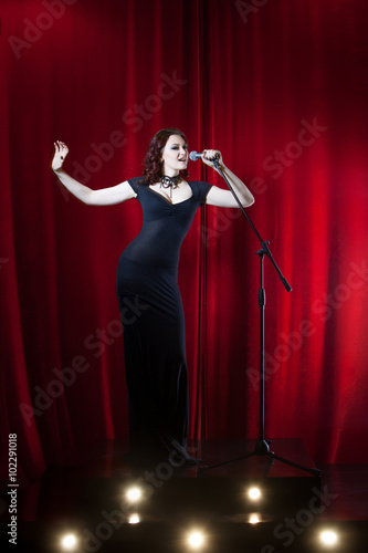 Beautiful Singing Girl on stage. Beauty Woman with Microphone. Red curtain  background