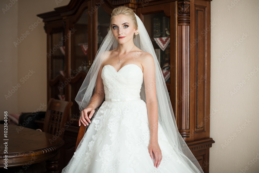 Elegant blonde bride in white wedding dress posing in hotel room