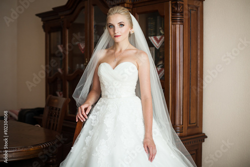 Elegant blonde bride in white wedding dress posing in hotel room