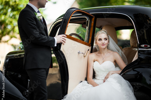 Gorgeous stylish blonde bride posing in retro black car with gro
