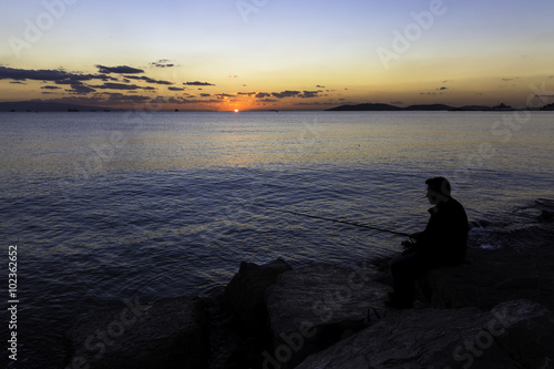 Scenic view of beautiful sunset with a fisherman silhouette sitting on the rocks near seaside © tolgaildun