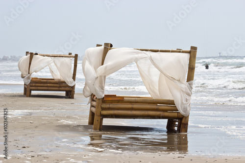 deux lits en bambou avec des voilages blancs posés au bord de l'eau sur la plage