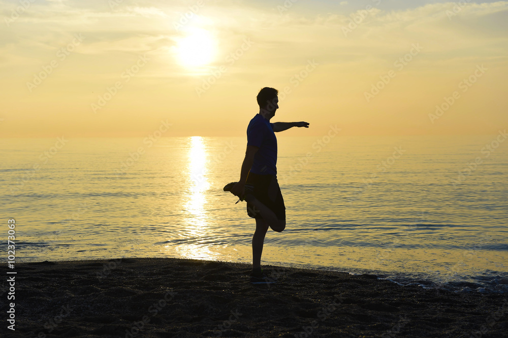 silhouette of young sport man stretching leg after running workout outdoors on beach at sunset