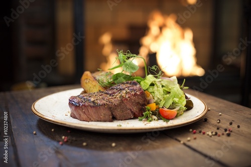 Delicious grilled stake and potatoes served on a wooden table, fireplace on background