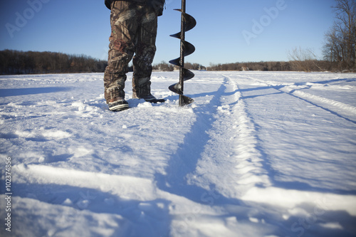 Canvas Print Ice fisherman drills hole with auger on lake in Minnesota