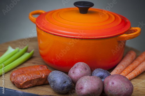 Orange enamel Dutch oven on cutting board with vegetables and cutlery on one side
