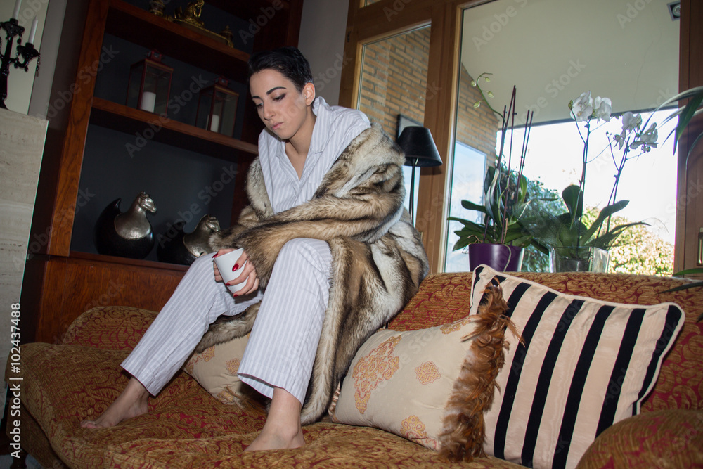 Caucasian girl with short hair sitting on the couch dressed in pajamas and fur coat. She is drinking coffee. 