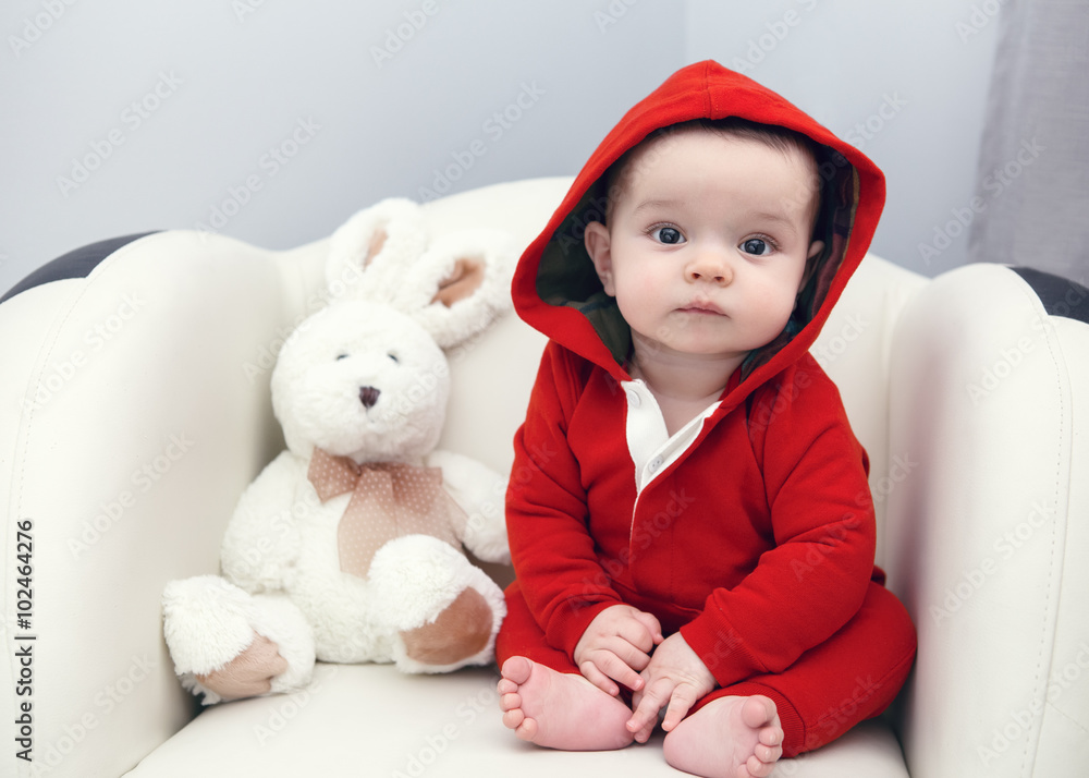 Portrait of cute adorable Caucasian smiling sad baby boy girl with black brown eyes in red hoodie shirt sitting in chair with toy looking directly in camera Portrait of cute adorable Caucasian smiling sad baby boy girl with black brown eyes in red hoodie shirt sitting in chair with toy looking directly in camera