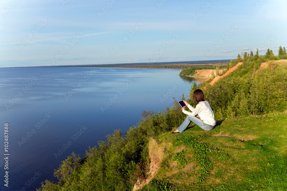 Naklejka premium Young woman sitting on the shore of the lake with tablet compute