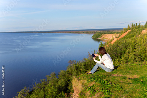 Young woman sitting on the shore of the lake with tablet compute