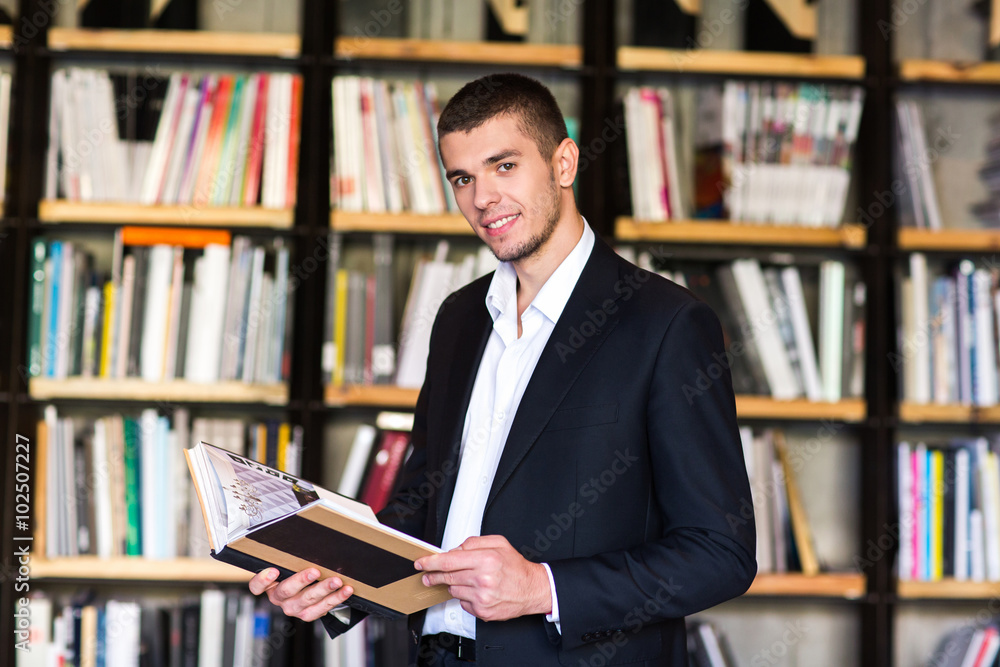 Handsome young man holding books and smiling while standing in library Handsome young man holding books and smiling while standing in library