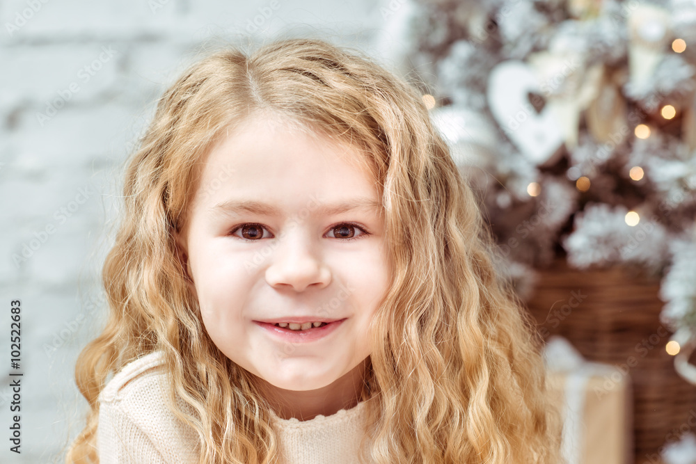 Pretty blond little girl sitting with long curly hair under the Christmas tree and smiling, closeup portrait Pretty blond little girl sitting with long curly hair under the Christmas tree and smiling, closeup portrait