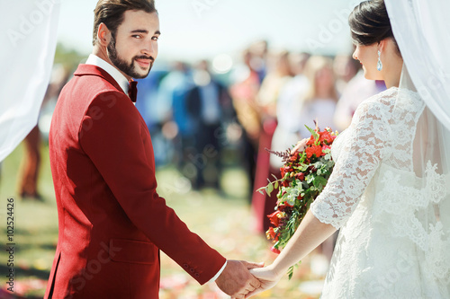 Groom pointedly looking at camera while standing in wedding ceremony arch with bride's hand on background of the crowd. New life concept. photo