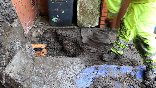 Plumber wearing bright reflective waterproof clothes replaced broken household waste drainage bottle gully outdoors and filling hole with dirt working in the rain photo