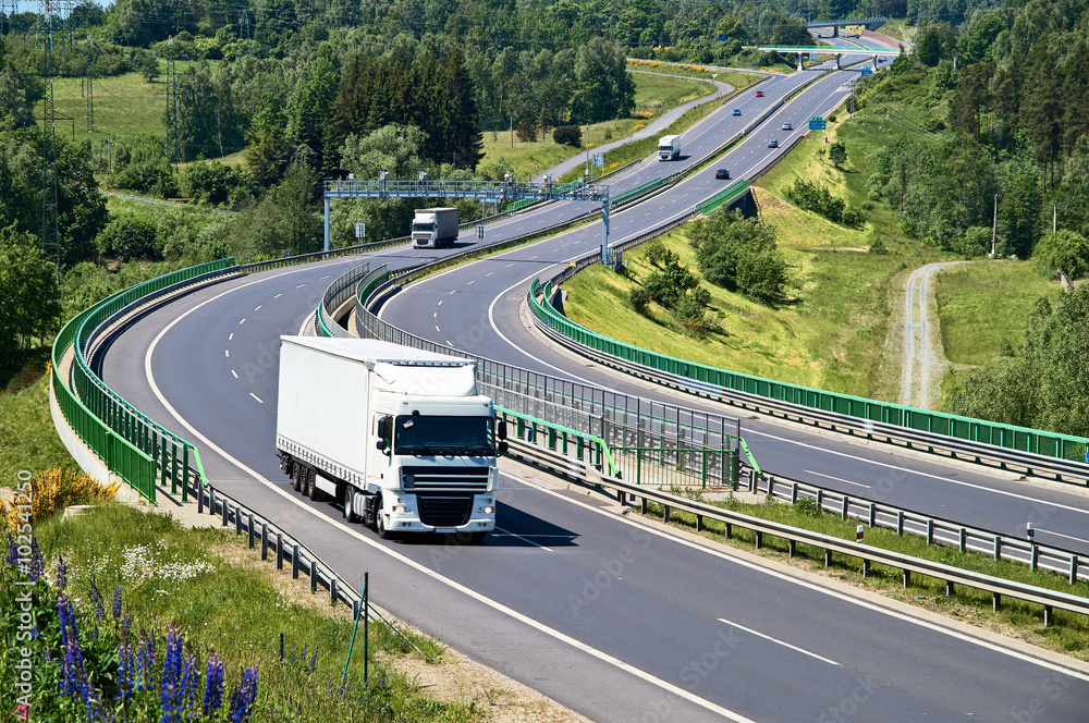 White trucks traveling on the asphalt highway with electronic toll gates in a wooded landscape. View from above. Sunny summer day.