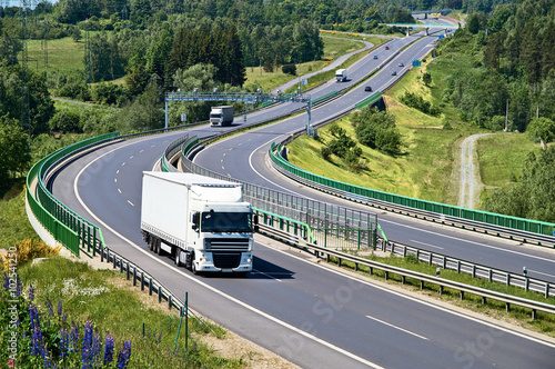 Obraz na płótnie White trucks traveling on the asphalt highway with electronic toll gates in a wooded landscape. View from above. Sunny summer day. w salonie